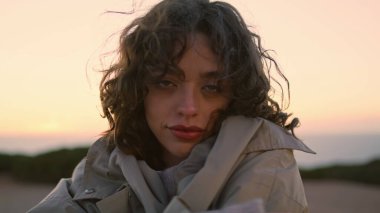 Romantic girl feeling cold adjusting coat closeup. Happy woman resting sunset. Attractive curly female looking camera getting warm hiding in clothes on seaside walk. Peaceful female posing outdoors