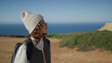 Girl walking cliff edge on sunny day. Happy tourist enjoying spring vacation going with backpack. Pretty woman traveler rest alone admire ocean landscape on autumn weekend. Tourism relaxation concept