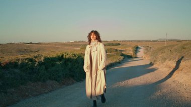 Walking girl enjoying evening sunlight on weekend. Single serene woman stretch feeling happy satisfied with life. Romantic attractive tourist going dusty road relax outdoors. Leisure serene lifestyle