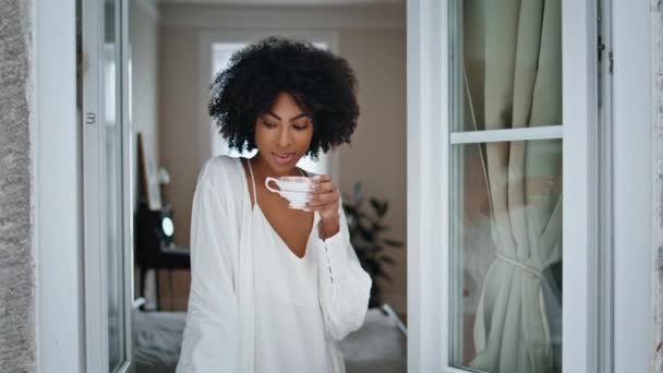 沉着的模特儿喝着咖啡靠窗的地方 快乐的非洲女人在家里呆着懒洋洋的早晨 松懈的女孩在客厅里享受着热饮 睡梦中的女士在周末休息 — 图库视频影像