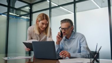 Modern ofiste işbirliği yapan gülümseyen iş adamları. Sakallı iş adamı telefonda konuşuyor. Mutlu kadın asistan dizüstü bilgisayarın yakın çekimini gösteren belge okuyor. Kabinede birlikte çalışan iş ortakları 