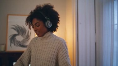 Kulaklıklı kadın akşam müziğinin keyfini çıkarıyor. Yakın plan gülümseyen bayan evde tek başına dans ediyor, dinleniyor. Modern kablosuz cihazda şarkı dinleyen Afro-Amerikalı neşeli kız. Tasasız yaşam tarzı konsepti