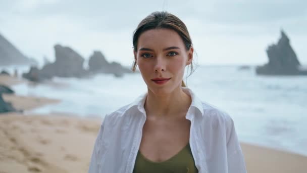 白いシャツを着て曇りのビーチに立っている若い女性が閉じます 波の前でカメラを見て魅力的な思慮深い少女の肖像画 Sereneシリアス美しいモデルが海岸でリラックス — ストック動画