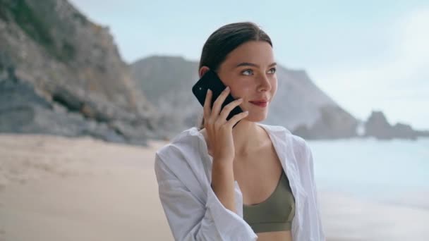 快乐美丽的女孩站在灰暗的沙滩上 用智能手机说话 在多云的海滨 一个迷人而轻松的女人打电话给我的肖像 在高山前面拿着手机的女士 — 图库视频影像