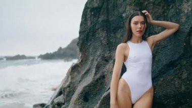 Kayalık plajda poz veren seksi kız yaz haftasonu kapalı. Taş uçurumdaki kameraya dayanan beyaz mayo giymiş çekici bir kadın. Güzel model plaj bulutlu gününde duruyor..