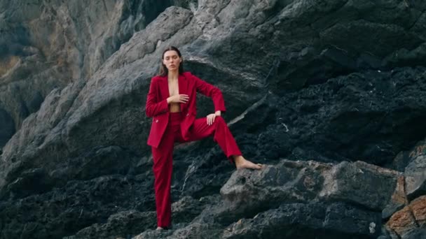 自信を持ってセクシーな女の子山の岩の上に豪華な赤いスーツ曇りの日を着てポーズ 石の岩の曇りの週末に立って深刻なスタイリッシュな女性 カメラを見て豪華なセクシーなモデル ファッションコンセプト — ストック動画