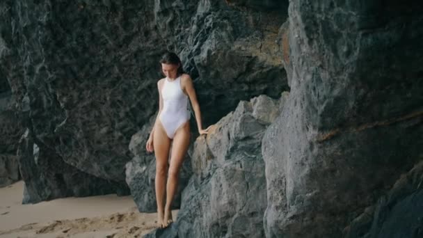 优雅漂亮的姑娘站在岩石前 在镜头前摆出一副很敏感的姿势 迷人的性感女人穿着白色泳衣靠在海滨悬崖上 瘦小华丽的女士在海滨自然中放松 — 图库视频影像
