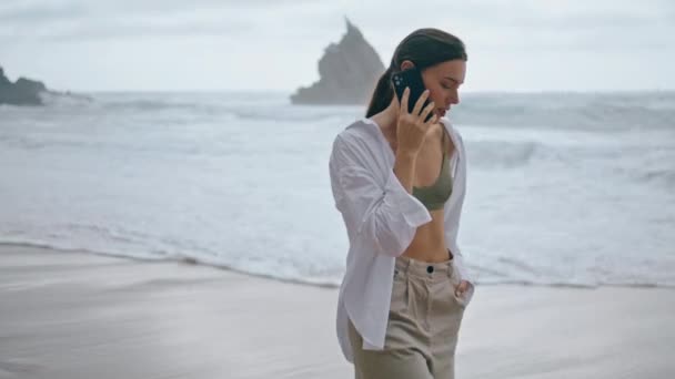 说电话的年轻姑娘松懈了 走近了泡沫般的海浪 一个人在多云的海滩上散步时 无忧无虑的迷人女人在讲电话 穿休闲装的沉着女士有电话交谈 — 图库视频影像