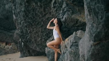 Taş kayaya yaslanmış sahilde poz veren seksi genç kadın. Deniz kıyısında beyaz mayo giyen çekici, şehvetli bir model. Uçurumun kenarındaki kumsalda duran muhteşem seksi kız. Güzellik konsepti.