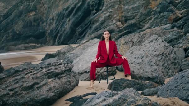 セクシーな赤いスーツを着て石の海岸にポーズをとる誘惑的なエレガントな女の子 椅子悲観的な性質に座って魅力的なグラマラスモデル 美しい曇りの海を探してスタイリッシュな若い女性 美の概念 — ストック動画