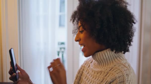 在家里用智能手机的积极女孩关门了 卷曲微笑的女士尝试新的手机应用程序在晚上的房子 非洲裔美国女人在客厅里读着令人愉快的信息 — 图库视频影像