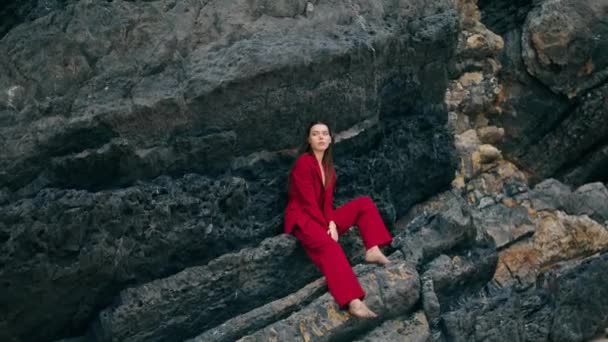 スタイリッシュな赤いスーツを着て岩の下に座って豪華なエレガントなモデル 美しい魅力的な女性の石の崖の曇りの日にポーズ セクシーな可愛い女の子の山の棚感の静けさにリラックス 美の概念 — ストック動画