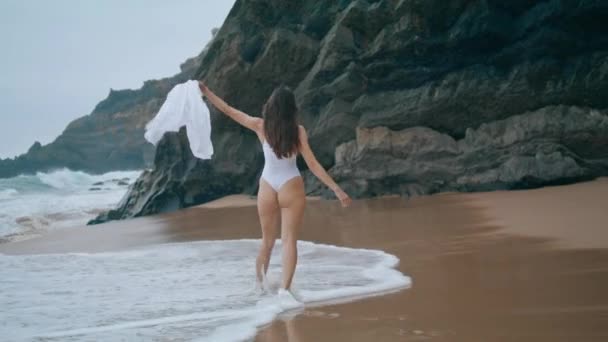 在多云的海滩上 快乐的女性游客享受着夏日的海浪 年轻迷人的女孩穿着性感的白色泳衣在海滨散步 一位无忧无虑的微笑女士站在高高的悬崖前 — 图库视频影像