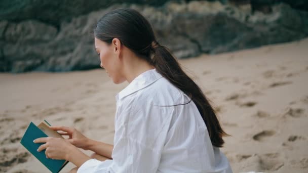 年轻女子穿着白衬衫坐在沙滩上看书 留著长发 专心致志的女孩手牵着文学 独自在海滨放松 迷人的女士在海滨享受平静的周末 — 图库视频影像