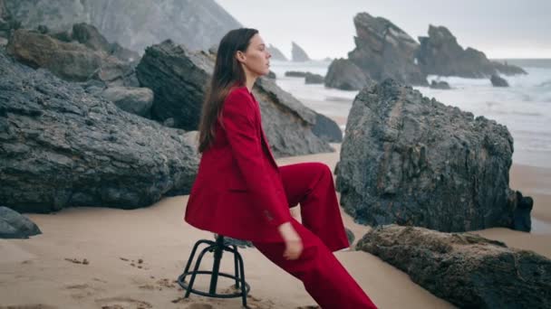 忧心忡忡的女人坐在岩石海滩上的椅子上 看着汹涌的大海 穿着红色雅致西服的迷人姑娘自信地看着岩石海岸 多云海岸上的性感模型 — 图库视频影像