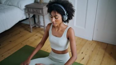 Yatak odasında bacaklarını açan rahat bir kadın. Kıvırcık saçlı esmer, lastik halıya meditasyon yaparken müzik dinliyor. Afrikalı sakin kadın sabah yüksek açıyla vücudunu ısıtıyor. Kız evde egzersiz yapıyor.