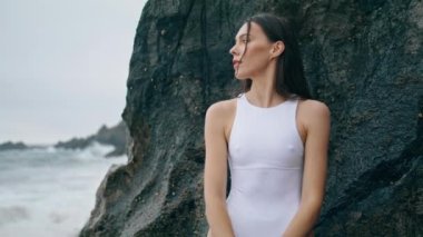 Güzel deniz manzarası seyreden şehvetli genç bir kadın kayaya yakın duruyor. Bulutlu sahilde duran beyaz seksi mayolu çekici nazik kız. Muhteşem genç model fırtınalı okyanus dalgalarını izliyor..
