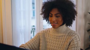 Evde bilgisayarla sohbet eden olumlu bir kız var. Kıvırcık gülümseyen bayan iş yerinde arkadaşına mesaj atıyor. Oturma odasındaki sosyal medyayı kullanan Afrikalı Amerikalı bir kadın.