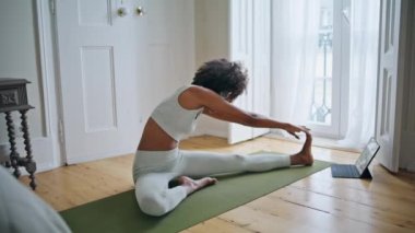 Yoga minderinde esnek model esneme. Kıvırcık saçlı spor antrenörü, Asya oturma halısı çalışıyor. Afro-Amerikalı kız tek başına tablet video eğitimi izliyor. Uyumlu yaşam tarzı kavramı 