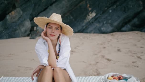 快乐宁静的女孩坐在岸上毛毯上 戴着宽大的草帽 无忧无虑的迷人女人微笑着在沙滩上放松地拍照 有魅力的年轻女士在海滨度假愉快 — 图库视频影像