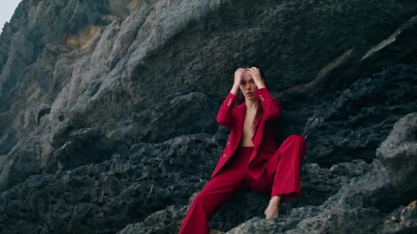 裸体でエレガントな赤いスーツを着た石の崖にポーズをとるセクシーなポーズの女性 カメラを自信を持って長い髪に触れるゴージャス本格的な女の子 美しいです魅力的な女性座っています下岩エッジ — ストック動画