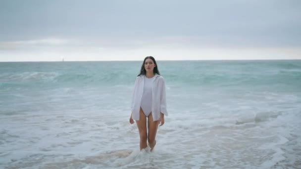穿着性感的白色泳衣在泡沫中穿行的宁静美丽的女人 长毛的年轻女士在沙滩上的阴天去沙滩 放松迷人的女孩在海滨享受平静忧郁的周末 — 图库视频影像