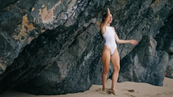 炎热的年轻女子站在沙滩上 手举到高高的岩石上 迷人的性感模特穿着白色优雅的泳衣 在石崖边的海滩上摆姿势 漂亮的长发姑娘在户外放松一下 — 图库视频影像