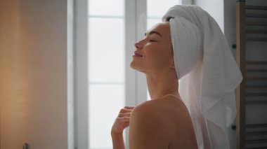 Calm girl cleaning neck in bathroom closeup. Confident woman holding cotton pad doing morning routine at modern apartment. Gorgeous lady with towel on head standing alone looking mirror at home