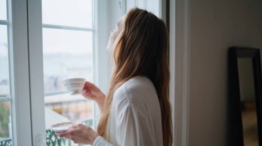 Satisfied lady enjoying cup of coffee at home closeup. Attractive woman drinking tea watching window at morning. Relaxed girl having rest looking city view at cozy apartment. Daily rituals concept