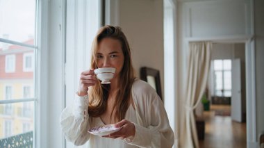 Positive girl drinking tea at home portrait. Happy lady holding saucer standing at window place close up. Smiling woman posing camera at morning apartment. Relaxed model sipping coffee at house