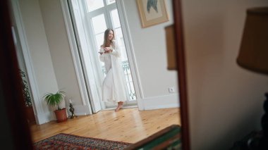 Dreaming model watching window at cozy interior. Mirror reflection of tender woman holding tea cup at light apartment. Positive girl drinking coffee standing at home. Carefree lady enjoying morning