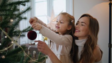 Evde Noel ağacı süsleyen sevimli bir kadın çocuğu. Gülümseyen anne kızını şefkatle kucaklıyor. Sevimli bir aile, bayramlarda dallara takılarak eğleniyor. Yeni yıl kutlaması 
