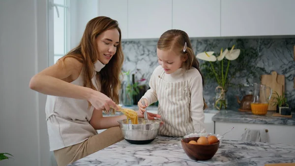 Loving mother teach daughter bake in kitchen. Friendly family cooking cake together. Single mother cleaning finger of baby girl at modern home. Little child having fun with cheerful woman indoors