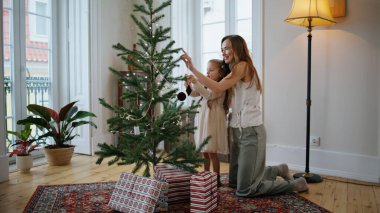 Evdeki Noel ağacını süsleyen neşeli anne kız. Gülümseyen anne çocuk oturma odasındaki Xmas ormanına oyuncak koyuyor. Sevimli aile yeni yıl kutlaması için hazırlanıyor. Kış tatili kavramı 