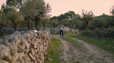 Tanned guy observing garden nature alone. Serious man spending time at olive trees plantation. Relaxed farm worker walking calmly at stone wall place. Male person crossing path at rural terrain 