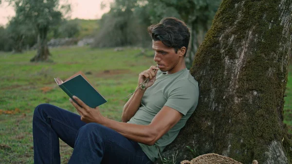 Focused man flipping book at evening park closeup. Relaxed garden worker reading favourite literature at rural landscape. Peaceful adult guy enjoying fresh air at summer nature zoom on. Break concept