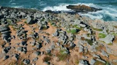 Güneşli, kayalık sahil sinematik hava manzarası. Mavi köpüklü okyanus dalgaları manzaralı kayalıklara çarpıyor. Fırtınalı deniz sörfünün insansız hava aracı sahile yavaş çekimde sıçrıyor..