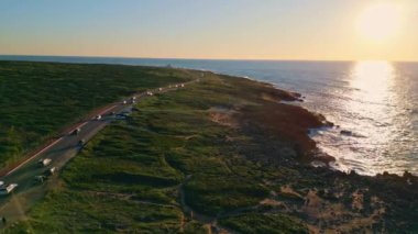 Yazın gün batımında, kayalık sahillerde sahil yolu. Deniz kıyısında ağır çekimde giden arabaların sinematik drone görüntüsü. İnanılmaz gün batımı dalgalı okyanus yüzeyine yansıyor. Turizm konsepti.