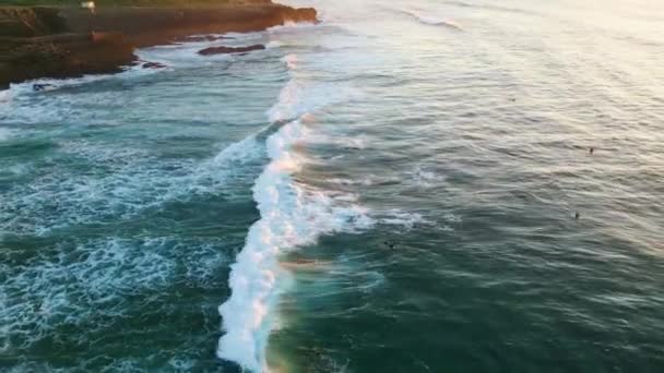 夏の夜に岩だらけの海岸を転がる深い青い海 波を待っているサーフボードに横たわっている未知のサーファー 日没の遅い動きで海岸線に走る空中風景の海泡 — ストック動画