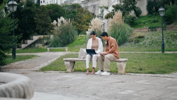大学生はベンチでタブレットを学ぶ 緑の公園地帯で働くタブコンピュータを使用して刺激的な若いビジネスカップル 屋外で話している文書をチェックする自信のある同僚 チームワークのコンセプト — ストック動画