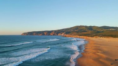 Panoramik deniz manzarası yaz günbatımı. Arkaplanda yeşil yamaçları olan kumlu sahilde hava manzaralı köpüklü okyanus dalgaları. Turkuaz deniz suyu, tropik deniz kıyılarını akşam gökyüzü altında yavaş çekimde yıkıyor.