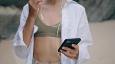 Bilinmeyen bir genç kız, kumsalda dikilmiş kulaklık takıyor ve beyaz gömlek giyiyor. Slim rahat kadın kulaklıkla akıllı telefondan müzik dinlemeye başlıyor. Güzel bayan mobil uygulamayı açıyor.