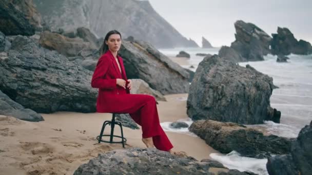 前の灰色の海の岩の多い曇りのビーチで椅子に座って官能的な若い女の子 曇った石の海岸で赤いエレガントなスーツを着たファッショナブルなグラマラスな女性 自然上のセクシーな自信モデルルッキングカメラ — ストック動画