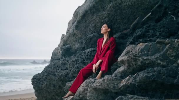 エレガントな赤いスーツで岩の棚の下に座っているリラックスしたスタイリッシュな女性 暗い劇的な海の景色を見て思慮深い若い女の子 秋の海岸で前の石の崖にポーズをとるファッショナブルな貴族の女性 — ストック動画