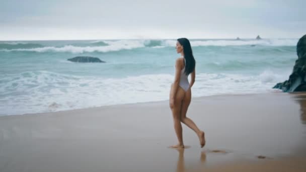 一个松懈的年轻女子在多云的暴风雨中去游泳 迷人的长发女孩穿着白色性感泳衣走在潮湿的海滩沙滩上 面对着泡沫般的海浪 美丽的女士在海滨休息 — 图库视频影像