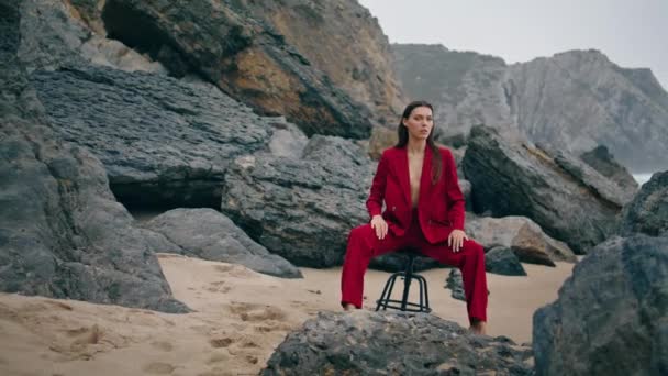 豪華なモデル岩の海岸悲観的な曇りの夜に椅子に座っている フロントドラマの嵐の海で赤いエレガントなスーツを着てポーズ官能的な美しい女性 誘惑的なスタイリッシュな女性は穏やかな夕暮れをお楽しみください — ストック動画