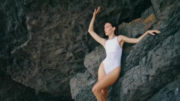 穿着白色雅致泳衣的性感年轻女子靠在海滩岩石边 迷人的性感女孩看着镜头 充满诱惑地站在石崖边 性感华丽的时装模特放松一下自然 — 图库视频影像