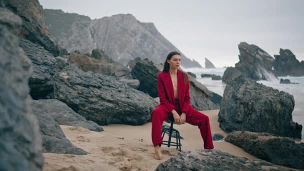 セクシーな赤いスーツを着て岩のビーチで椅子に座って豪華な女性 岩の崖の海岸曇りの日にポーズを裸の体にジャケットと魅惑的な若い女の子 嵐の海の景色を見る高級モデル — ストック動画