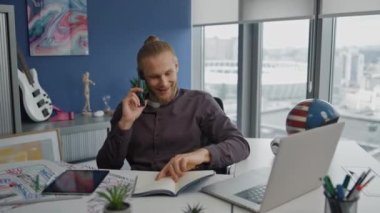 Ofiste akıllı telefondan konuşan gülümseyen bir adam. Bilgisayar masasında defter izlerken telefonda mutlu başlangıç konuşmaları. Modern uzman pencere işyerindeki iş ortağına ayrıntıları açıklıyor