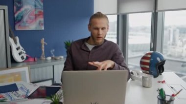 Hareketli adam modern ofisten dizüstü bilgisayarı arıyor. Panoramik pencere odasında yetenekli bir tasarımcı sanal konferans veriyor. Uzaktaki bilgisayar toplantısında ortaklarla ciddi bir video oluşturma sohbeti